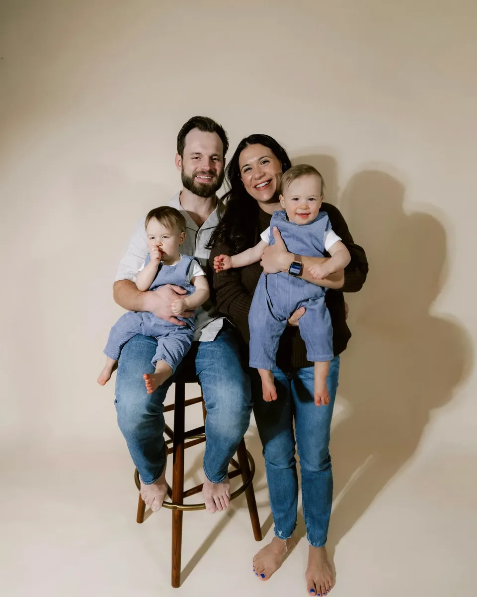 Família de quatro posando em estúdio com fundo claro, com dois bebês no colo